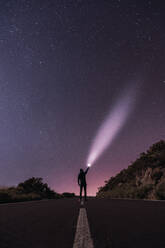 Rückenansicht eines Mannes, der auf einer leeren Fahrbahn steht und mit einer Taschenlampe in den dunklen Sternenhimmel leuchtet - ADSF09872