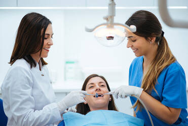 Side view of careful smiling dentist and assistant making examination of oral cavity with mirror and salivary tube - ADSF09881
