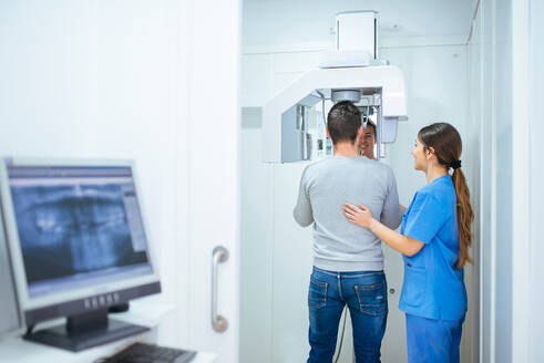 Rückenansicht eines Kunden, der mit Hilfe einer sorgfältigen Assistentin in blauer Uniform in einem zahnärztlichen Kabinett mit Monitor und Ausrüstung geröntgt wird - ADSF09885