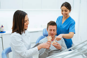 Begeisterter Patient auf einem Stuhl, der eine Prothese hält, und Zahnarzt mit Assistentin in Uniform, die die Zahnstruktur erklärt - ADSF09886