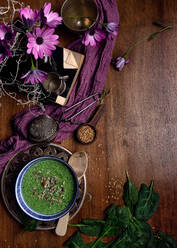 Von oben Keramikschale mit frischem grünem Smoothie mit Kürbiskernen und Sesam in Komposition mit lila zartem Stoff und Strauß frischer violetter Gänseblümchen in Vase auf braunem Holztisch platziert - ADSF09895