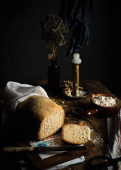 Loaf of homemade bread on cutting board and fresh curd cheese placed on kitchen table with candlestick and vase with plant - ADSF09907