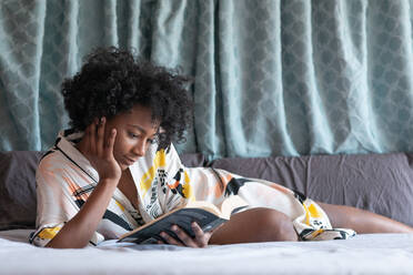 Content African American female in silk robe lying on comfortable bed and enjoying novel while leaning on hand - ADSF09928