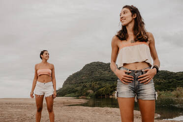 Zwei schlanke junge Frauen in Shorts und BHs lächelnd und Blick auf verschiedenen Seiten, während stehend auf sandigen Ufer gegen bewölkten grauen Himmel - ADSF10008