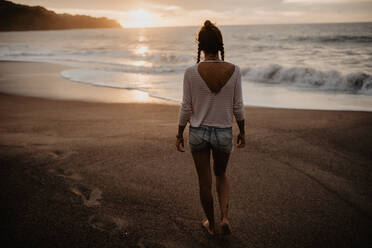 Rückenansicht einer jungen Frau in Freizeitkleidung, die am Sandstrand in Richtung stürmisches Meer bei Sonnenuntergang in der Natur spazieren geht - ADSF10010