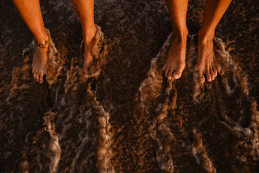 Barefoot legs of unrecognizable women standing on wet shore near splashing sea in evening - ADSF10012