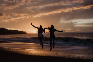 Silhouettes of anonymous female friends holding hands and leaping towards waving sea against cloudy sunset sky - ADSF10013