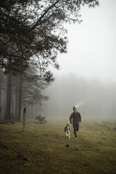 Confident male runner in sportswear training in misty wood with dog while jogging and looking away - ADSF10018