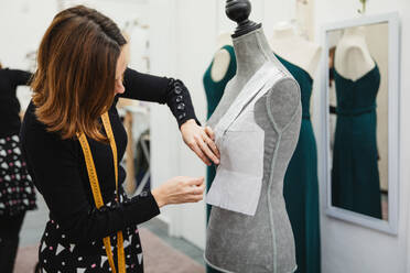 Anonymous woman with tape measure attaching paper cutout to mannequin with pin while making clothes in professional workshop - ADSF10022