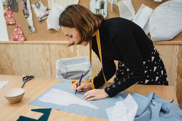 Erwachsene Frau, die ein Maßband benutzt, um ein Kleidungsstück auf dem Tisch zu messen, während sie in einer professionellen Schneiderei arbeitet - ADSF10025