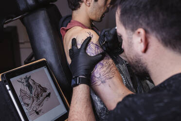Konzentrierter Künstler bei der Tätowierung auf der Schulter eines männlichen Kunden, während er einer Skizze auf einem Tablet in einer Werkstatt folgt - ADSF10035