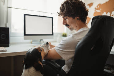 Seitenansicht eines ernsthaften männlichen Unternehmers, der auf einem Bürostuhl sitzt und einen niedlichen Hund bei der Fernarbeit betrachtet - ADSF10036