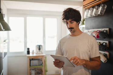 Serious male in casual clothes standing in modern kitchen and browsing tablet - ADSF10037