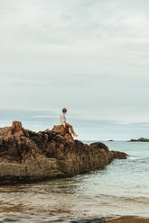 Lonely tourist sitting on rocky coast against tranquil sea water under gray sky - ADSF10041