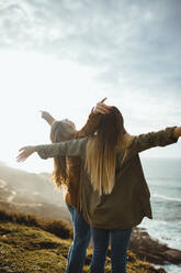 Seitenansicht von jungen Frauen im Gegenlicht, die die Hände heben und die Haare über den Köpfen halten, während sie zusammen am Meer stehen und die frische Brise und die Freiheit genießen - ADSF10046