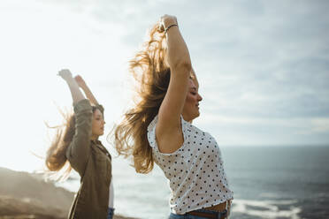 Side view of young women in back lit raising hands and holding hair over heads while standing together at seaside and enjoying fresh breeze and freedom - ADSF10047