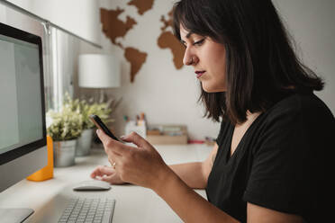 Seitenansicht der Ernte konzentriert Frau mit dunklen Haaren Messaging auf Handy beim Sitzen mit Desktop-Computer im Büro in Paris - ADSF10048