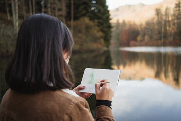 Rückenansicht einer Künstlerin, die neben einem schönen See mit Wald im Herbst steht und in ein Notizbuch zeichnet - ADSF10059