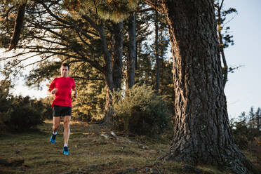 Aktiver gesunder erwachsener Mann in rotem Hemd und schwarzen Shorts beim Joggen auf einer mittelschweren Straße im Wald an einem sonnigen Herbsttag mit blauem Wolkenhimmel im Hintergrund - ADSF10067