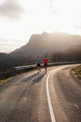 Active healthy male joggers running together on curved asphalt road with sunlight from behind mountain in background - ADSF10069