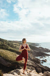 Frau in roter Sportkleidung, die trainiert und eine Baum-Yoga-Pose am leeren Felsstrand übt - ADSF10078