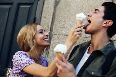 Side view of cheerful young attractive woman feeding boyfriend by cornet of ice cream outdoors - ADSF10090