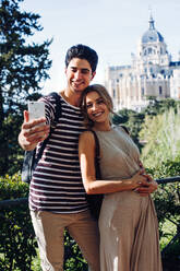 Young handsome man taking photo with girlfriend while walking in beautiful garden on background of historical building - ADSF10092