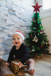 Glückliches Baby in Weihnachtsmannmütze und Pyjama, das mit einem Geschenk spielt, während es auf dem Boden in der Nähe des Weihnachtsbaums sitzt - ADSF10097