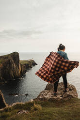 Rückenansicht einer einsamen Frau mit kariertem Plaid, die auf einem Klippenrand steht und gegen eine unscharfe Felswand und eine erstaunliche Meereslandschaft an einem windigen Tag in Schottland hinunterblickt - ADSF10109