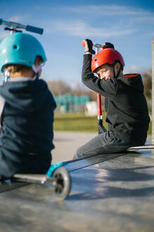 Side view of cheerful teen boy in helmet with kick scooter talking to unrecognizable friend while sitting together on ramp in skate park in sunny spring day - ADSF10145