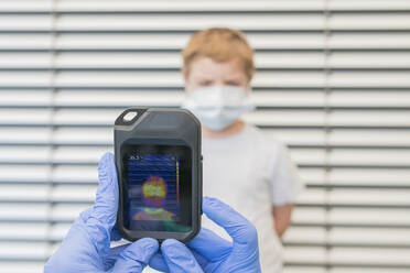Unbekannter Sanitäter in Schutzhandschuhen und Uniform bei der Verwendung einer Wärmebildkamera zur Überprüfung der Temperatur eines Kindes auf der Straße während des Ausbruchs des Coronavirus - ADSF10149