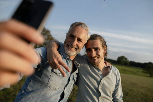 Glücklicher Vater mit erwachsenem Sohn macht ein Selfie auf einer Wiese auf dem Lande - KNSF08305