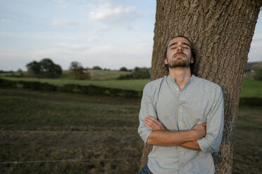 Relaxed young man leaning against a tree trunk in the countryside - KNSF08315