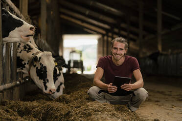 Porträt eines lächelnden jungen Landwirts mit Tablet im Kuhstall auf einem Bauernhof - KNSF08328