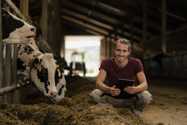 Portrait of a smiling young farmer with tablet at cow house on a farm - KNSF08328