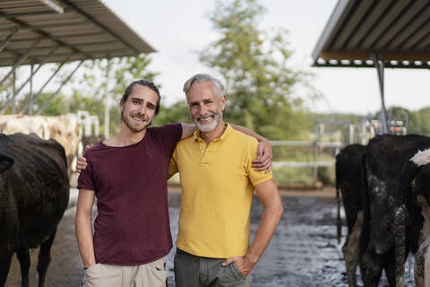 Portrait of happy mature farmer and adult son embracing at cow house on a farm stock photo