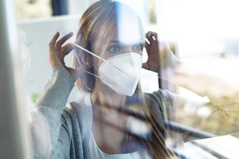 Porträt einer jungen Frau, die hinter einer Fensterscheibe eine Schutzmaske aufsetzt - JSRF00981