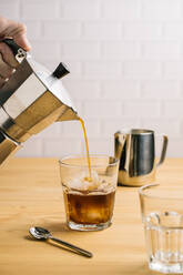 Crop person pouring coffee from coffeemaker into stylish glass with ice on wooden counter in cafe - ADSF10152