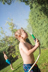 Little kid in swimwear splashing water from garden hose at himself - ADSF10159