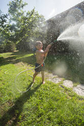 Kleines lachendes Kind in Shorts und mit nackten Füßen, das Wasser aus dem Gartenschlauch spritzt - ADSF10162