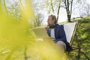 Nachdenklicher Geschäftsmann schaut weg, während er an einem sonnigen Tag mit seinem Laptop im Park sitzt - JOSEF01424