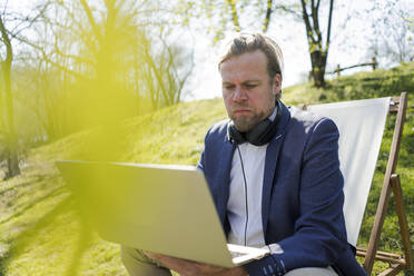 Reifer männlicher Unternehmer, der einen Laptop benutzt, während er im Sommer auf einem Stuhl im Park sitzt - JOSEF01425