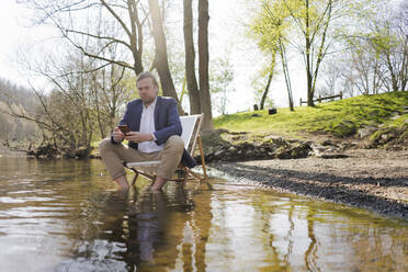 Männlicher Berufstätiger, der sein Smartphone hält, während er auf einem Stuhl am Flussufer vor Bäumen im Park sitzt - JOSEF01428