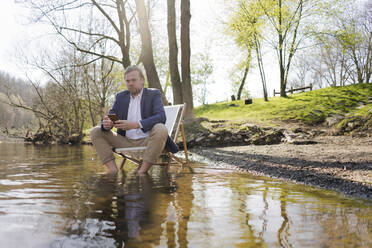 Male professional holding smart phone while sitting on chair at riverbank against trees at park - JOSEF01428