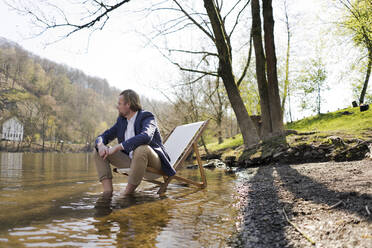 Nachdenklicher männlicher Berufstätiger, der auf einem Stuhl am Flussufer vor Bäumen im Park sitzt und wegschaut - JOSEF01430