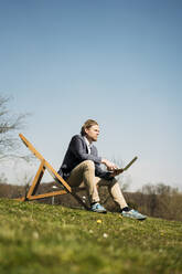 Oberflächenansicht eines männlichen Unternehmers, der wegschaut, während er mit einem Laptop auf einem Stuhl im Park an einem sonnigen Sommertag sitzt - JOSEF01431