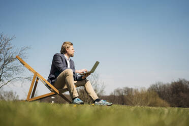 Oberflächenansicht eines Geschäftsmannes, der wegschaut, während er mit seinem Laptop auf einem Stuhl im Park an einem sonnigen Sommertag sitzt - JOSEF01432