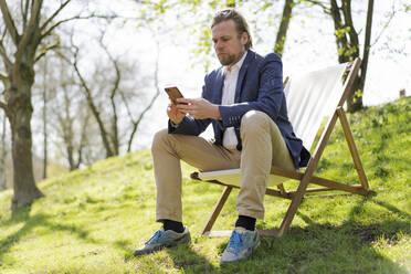 Männlicher Unternehmer, der sein Smartphone benutzt, während er auf einem Stuhl im Park sitzt - JOSEF01441