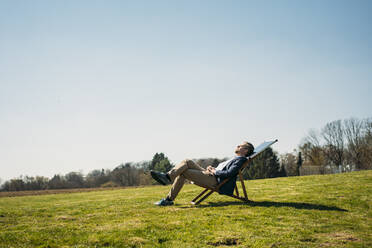 Mature male entrepreneur relaxing on chair with solar panel at park during sunny day - JOSEF01447