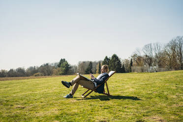 Mature male professional using smart phone while sitting on chair with solar panel at park during sunny day - JOSEF01448
